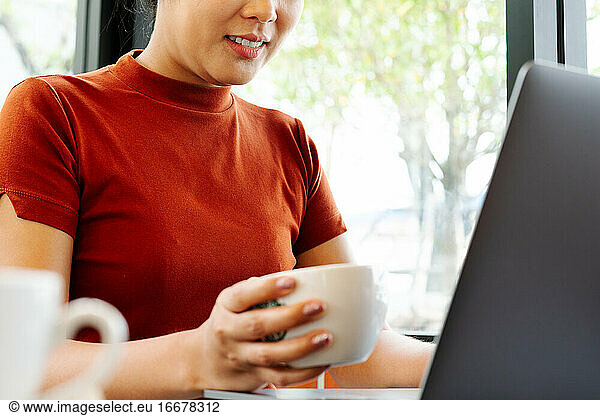 Asia woman hold cup of coffee while typing on laptop keyboard. W