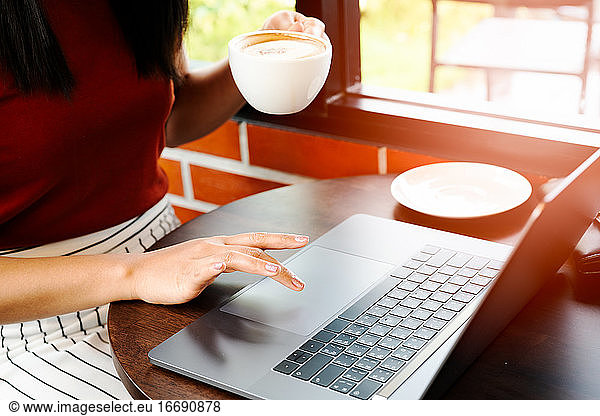 Asia woman hold cup of coffee while typing on laptop keyboard. W