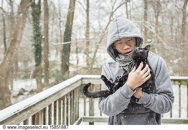 Asian boy lovingly takes curious black cat outside on a snowy day