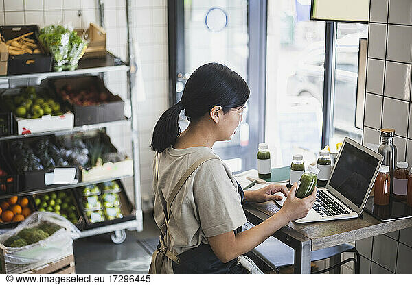 Asian female entrepreneur using laptop while holding juice bottle at store