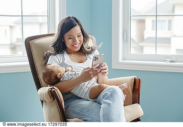 Asian mother holding newborn infant baby son and chat on smartphone