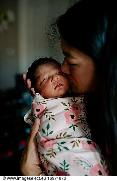 Asian mother kisses cheek of newborn baby swaddled in blanket