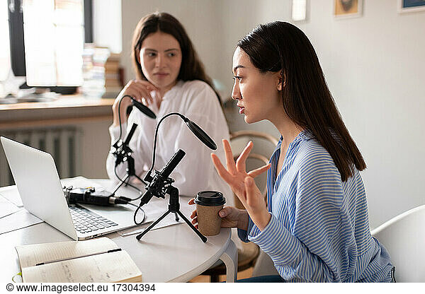 Asian woman with coffee to go speaking into microphone during podcast with friend