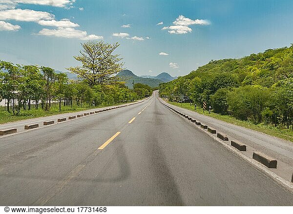 Asphalt road surrounded by greenery on a sunny day  Long asphalt road leading to a mountain with blue sky  Beautiful road on a sunny day leading to some mountains