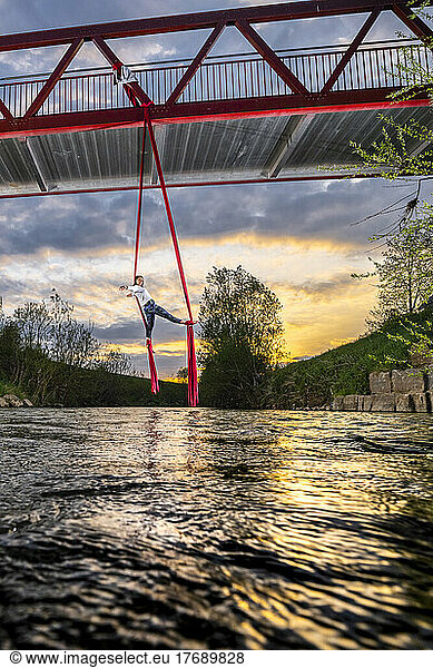 Athlete exercising aerial acrobatic hanging from bridge over Rems river at sunset
