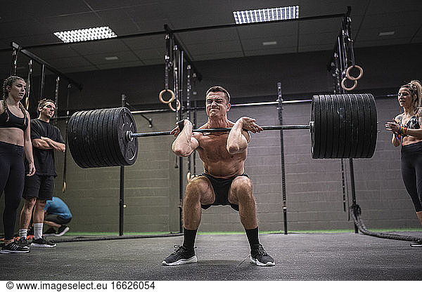 Athleten bejubeln einen Mann  der in der Hocke sitzt und eine Langhantel im Fitnessstudio aufhebt