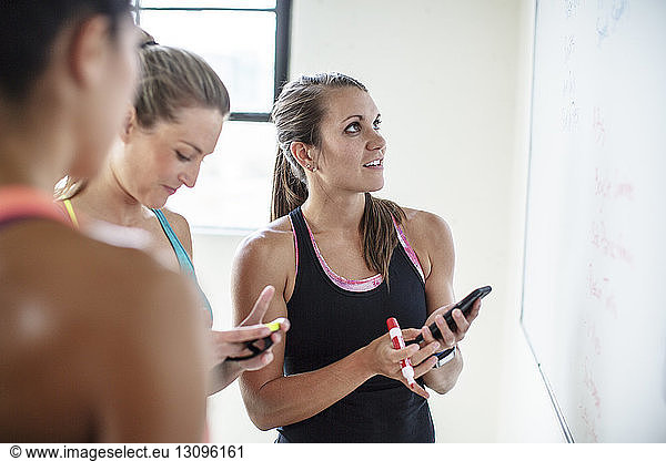 Athletes using mobile phone while standing with friend by whiteboard in gym