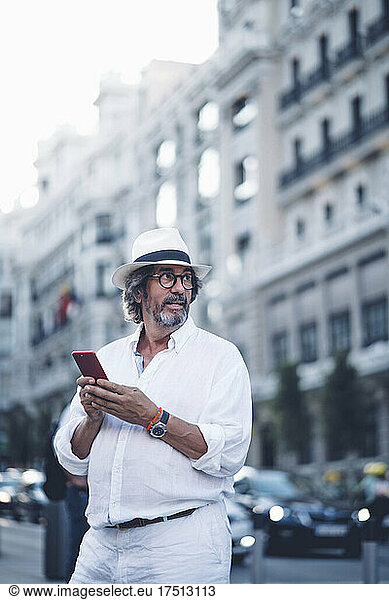 Attractive senior man with white clothing and hat looking away while holding smart phone in street of Madrid
