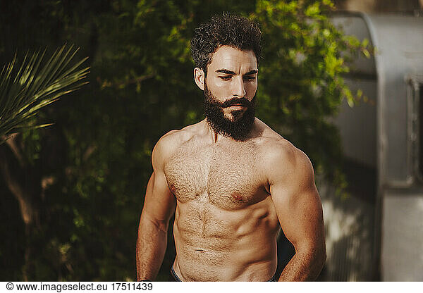 Attractive shirtless man standing in front of shrubs