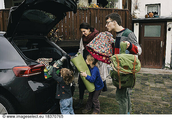 Aufgeregte Familie beim Einladen von Picknickutensilien in den Kofferraum eines Elektroautos vor dem Haus