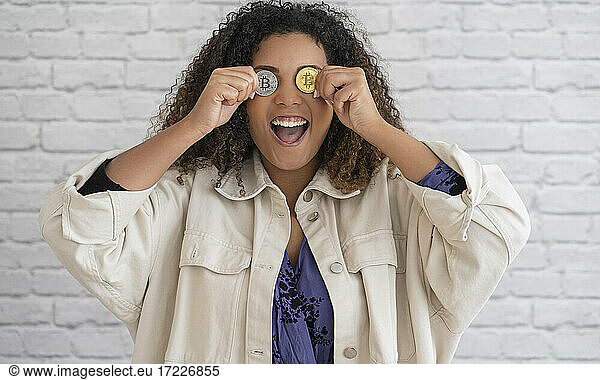 Aufgeregte lockige Frau  die ihre Augen mit gold- und silberfarbenen Bitcoins vor einer Wand bedeckt