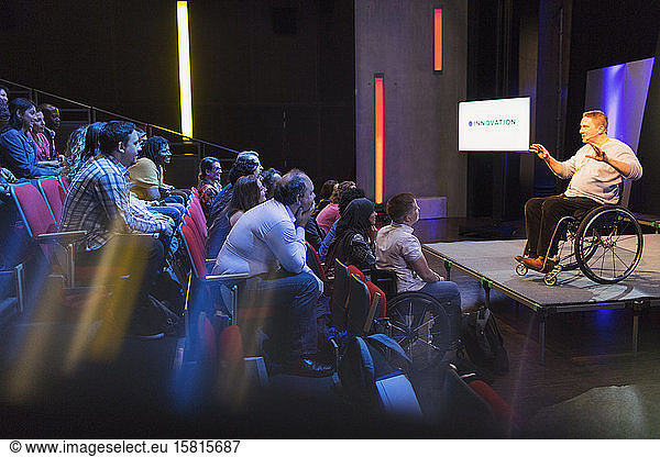 Aufmerksames Publikum hört einer Rednerin im Rollstuhl auf der Bühne zu