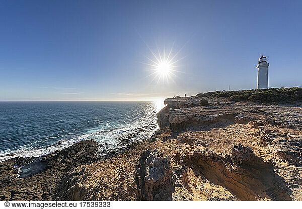 Australia  Victoria  Sun shining over rough coast of Cape Nelson State Park with Cape Nelson Lighthouse in background