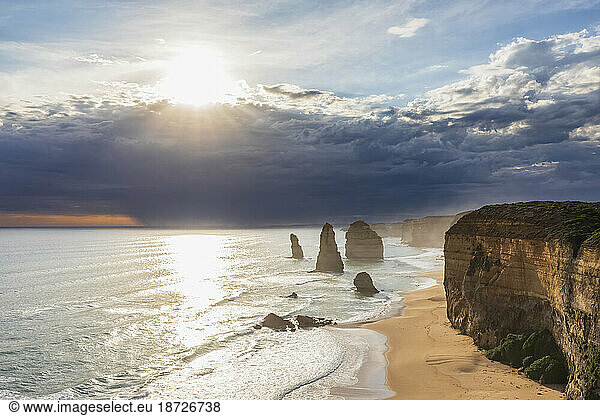 Australia  Victoria  View of sun shining through storm clouds over sandy beach in Port Campbell National Park with Twelve Apostles in background