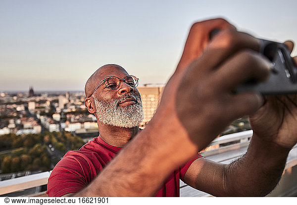 Bärtiger reifer Mann  der sein Smartphone benutzt  um den Sonnenuntergang von einem Hausdach in der Stadt zu fotografieren