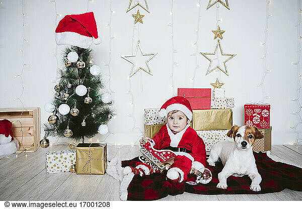 Baby boy in Santa Claus costume playing while sitting by dog at home during Christmas