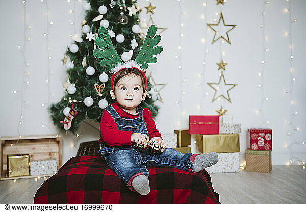 Baby boy wearing horned headband sitting against Christmas tree at home during Christmas