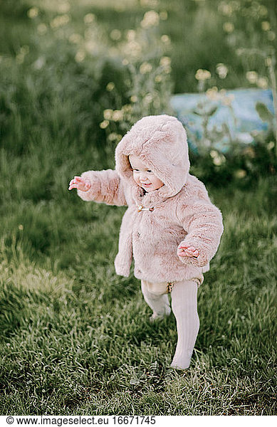 Baby girl first steps in pink fur coat outside on grass
