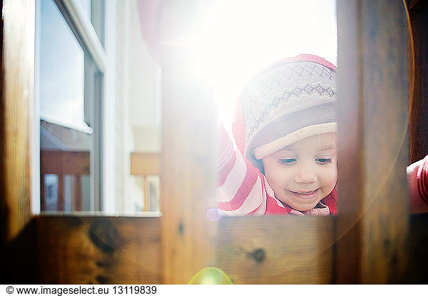 Baby girl in hooded jacket seen through wooden railing during sunny day
