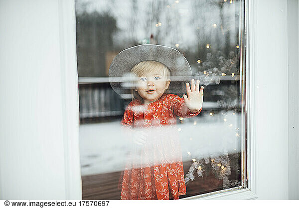 Baby girl standing and looking out window with hat by Christmas tree