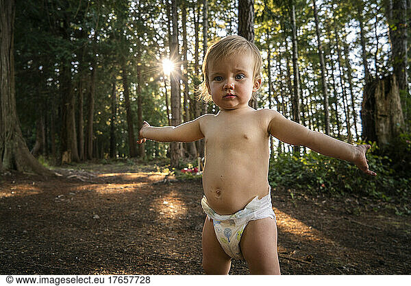 Baby in diaper stands with arms outstretched in forest