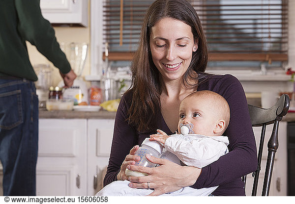Baby in mother's arm with pacifier and father making lunch