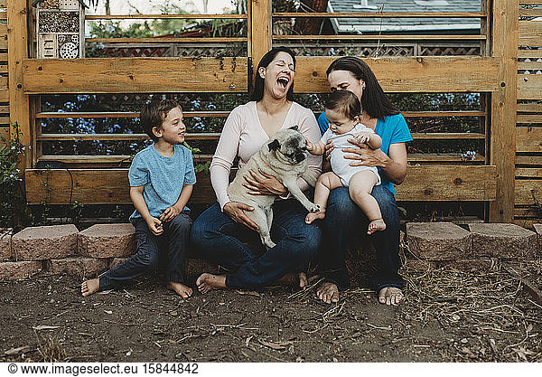 Baby touching pet pug with laughing barefoot two-mom family