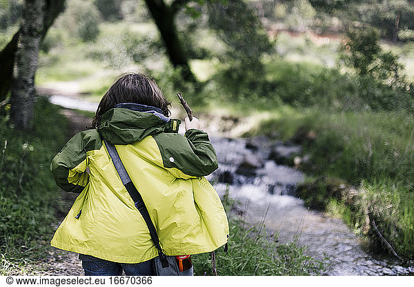 Back of kid hiking with green raining coat and stick in natural park