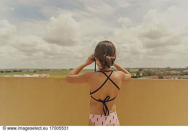 Back view of girl in swimsuit standing on roof terrace looking through binoculars