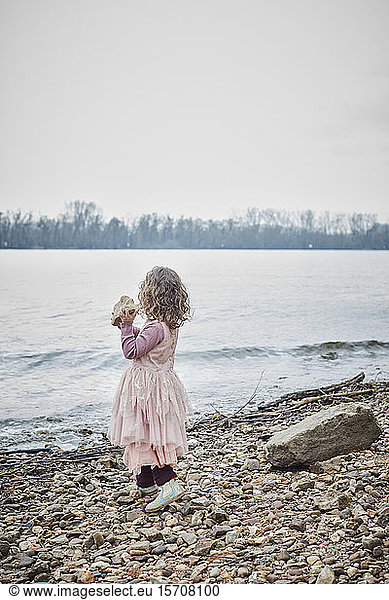 Back view of little girl wearing pink fancy dress costume standing at riverside holding a stone