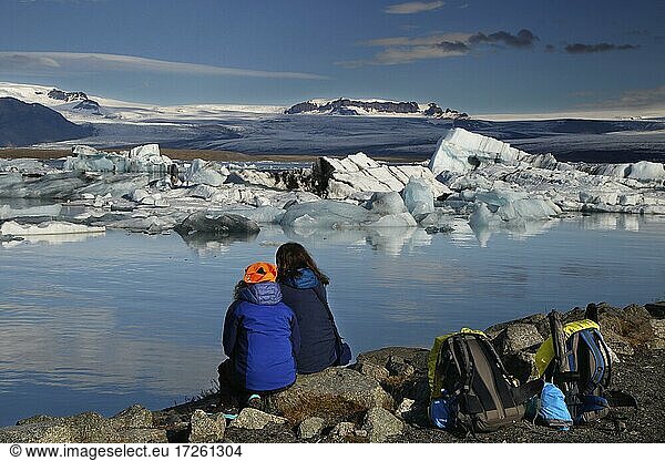 Back view of two tourists with backpacks at the water  icebergs  floating ice chunks  glacier ice  glacier  calving glacier  glacier lagoon  glacier lake  glacier lagoon Jökulsárlon  Vatnajökull glacier  south coast  Iceland  Europe