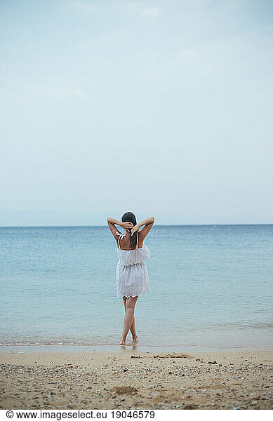 Back view of woman in white summer dress standing on sandy beach
