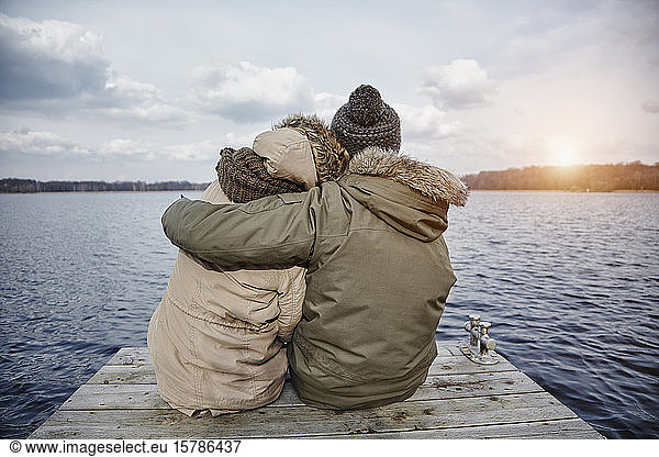 Back view of young couple in love sitting on a jetty looking at lake in winter