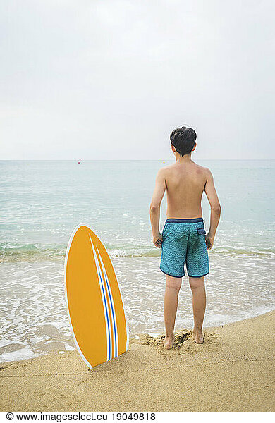 Back view of young teen with surfboard standing at seashore in a bright day