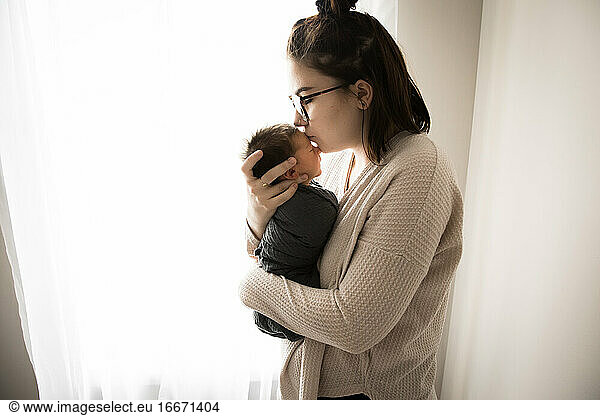 Backlit Gen Z Hipster Mom Kisses Newborn Baby in Front of Window