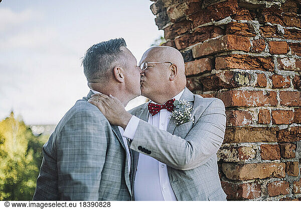 Bald gay man kissing groom on mouth by brick wall at wedding ceremony