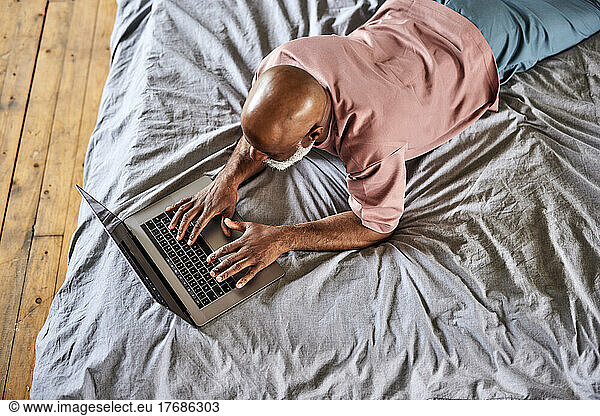 Bald man typing on laptop lying on bed at home