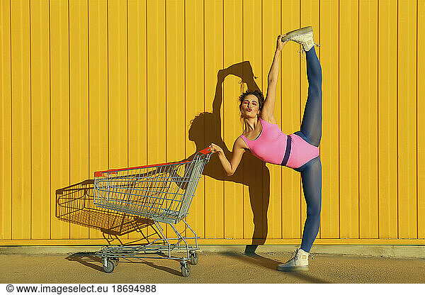 Ballet dancer doing vertical split by shopping cart in front of yellow wall