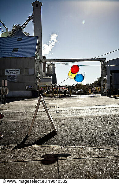 Balloons tied to a sign with a factory in the distance.
