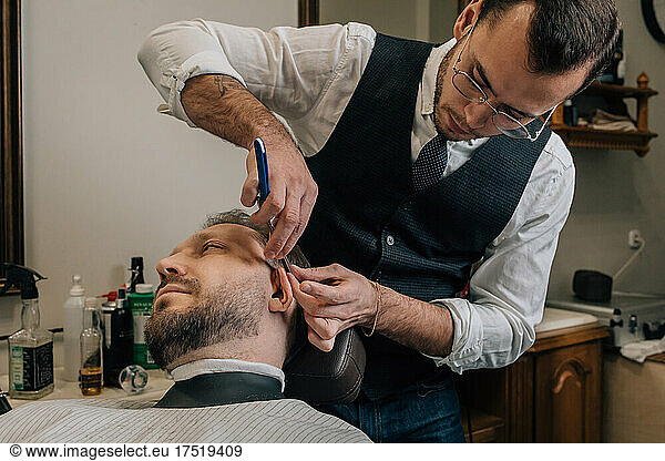 Barber hairdresser cuts facial hair with razor.