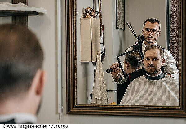 Barber shows client haircut from behind in reflection of mirror.