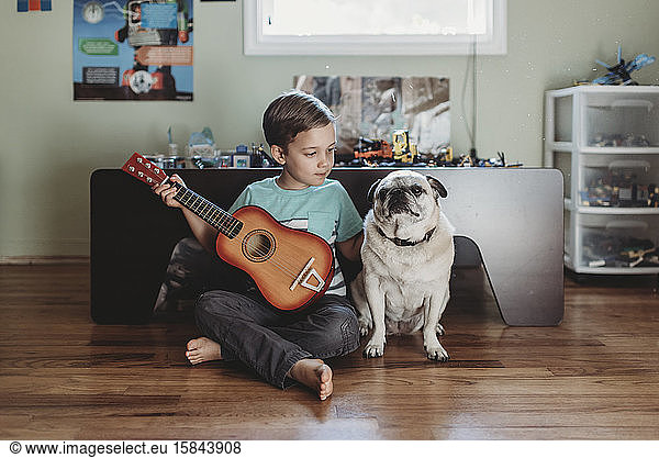 Barefoot boy holding guitar sitting next to pet Pug on hardwood floor