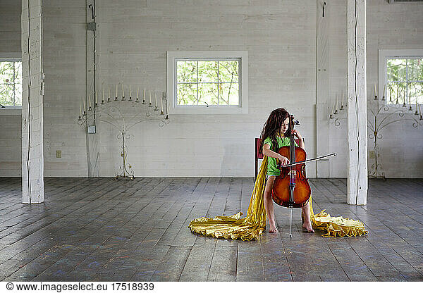 Barefoot girl in wig and gold cape plays cello in a large empty barn