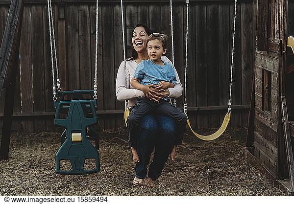 Barefoot laughing mom and young son on swing in backyard