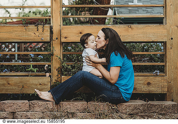 Barefoot mom kissing baby girl while sitting on landscape rocks