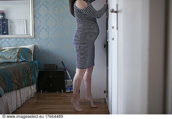 Barefoot pregnant woman in dress at closet in bedroom