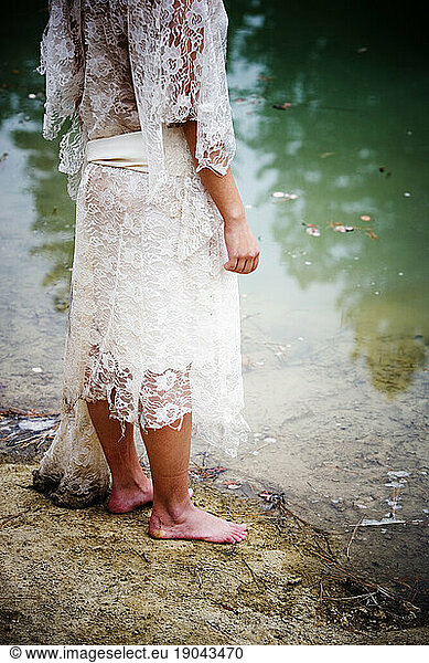 Barefoot woman in a white dress standing alone at the water's edge.