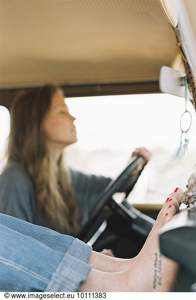 Barefoot woman resting her feet on the dashboard of a 4x4 a tattoo on her right foot another woman driving.