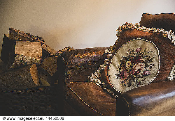 Basket of firewood next to vintage tan leather armchair with embroidered cushion with floral pattern.