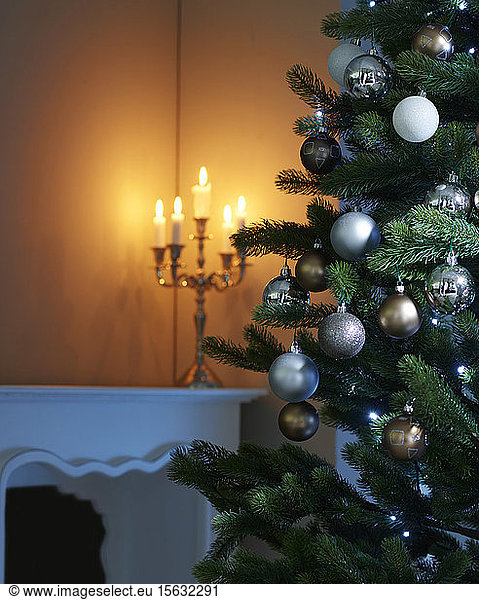 Baubles hanging on Christmas tree with candles burning in background at home